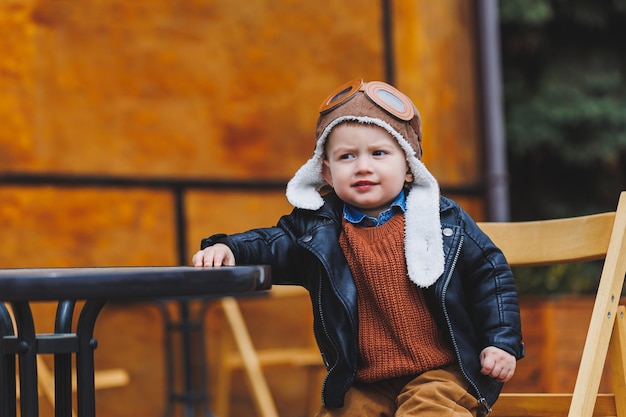 Stijlvolle jongen van 3 jaar oud in een leren jas en bruine broek zit op het terras bij het café Modern kind Kindermode Gelukkig kind