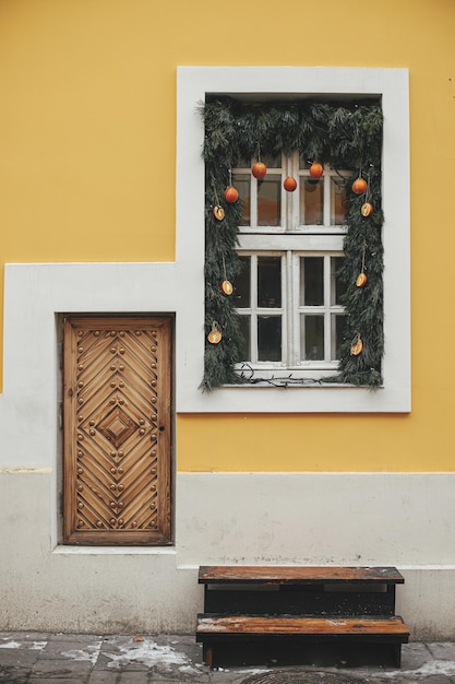 Stijlvolle kersttakken met sinaasappels en mandarijnen op wit raam van geel gebouw Modern eenvoudig eco-kerstdecor in stadsstraat Wintervakantie in Europa Vrolijk kerstfeest