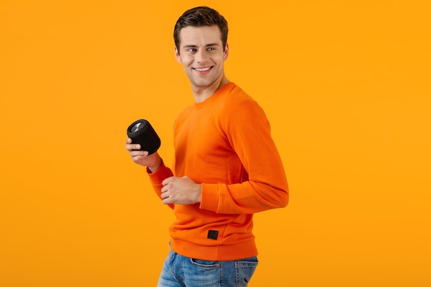 Stijlvolle lachende jonge man in oranje trui met draadloze luidspreker gelukkig luisteren naar muziek