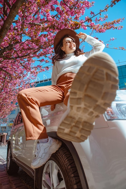 Stijlvolle lachende mooie vrouw staande in de buurt van auto bloeiende sakura boom op achtergrond