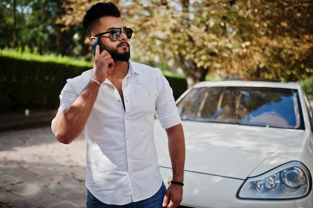 Stijlvolle lange arabische man model in wit overhemd jeans en zonnebril poseerde op straat van stad baard rijke aantrekkelijke arabische man tegen witte suv auto spreken op mobiele telefoon