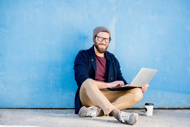 Stijlvolle man gekleed casual in trui en hoed werken met laptop zittend in de buurt van de blauwe muur achtergrond