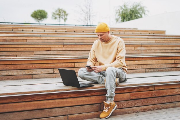 Stijlvolle man hipster in gele hoed freelancer werkt op laptop in het stadspark op straat