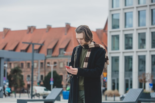 Stijlvolle man in sjaal en jas houdt mobiele telefoon in een hand in de straat van wroclaw, polen