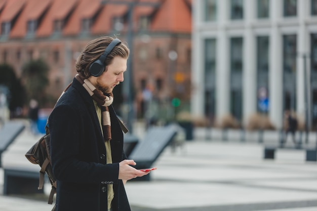 Stijlvolle man in sjaal en jas luistert naar muziek op streamingdienst met behulp van een mobiele telefoon in de straat van Wroclaw, Polen