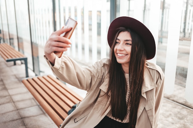 Stijlvolle meisje in hoed maakt selfie zittend op een openbare bushalte