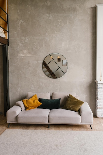 Stijlvolle moderne grijze bank met kussens in de woonkamer met grijze muren en een ronde spiegel in Scandinavische stijl