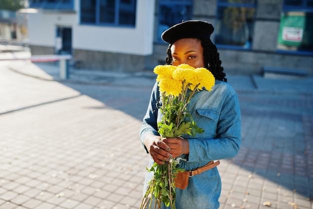 Stijlvolle modieuze Afro-Amerikaanse vrouwen in spijkerbroek dragen en zwarte baret met gele bloemen boeket buiten geposeerd in zonnige dag tegen blauw modern gebouw