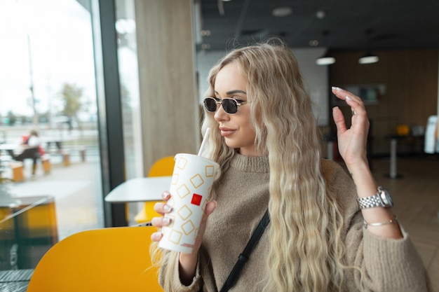 Stijlvolle mooie jonge blonde vrouw met krullende vintage zonnebril in een krullend kapsel in een mode beige trui zit in een café en houdt een papieren kopje cola vast