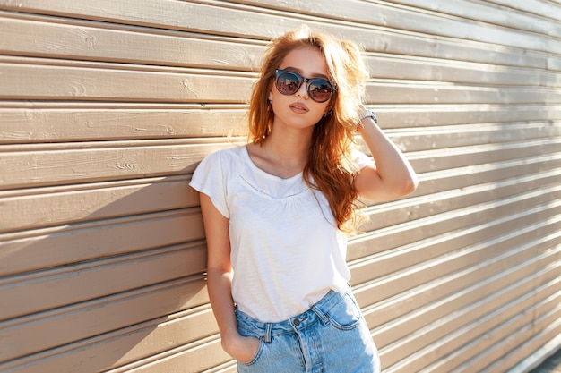 Stijlvolle mooie jonge model vrouw met zonnebril in een wit T-shirt en blauwe vintage jeans in de buurt van een houten muur bij zonsondergang