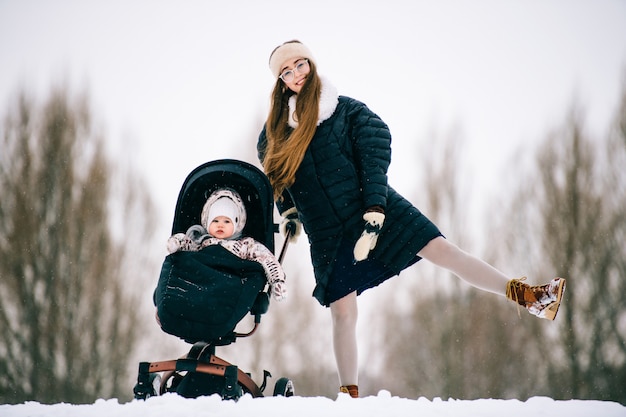 Stijlvolle mooie jonge moeder veel plezier samen met mooie kind zit in kinderwagen buiten in de winter. het gelukkige vrolijke vrouw en zuigelingsdochter spelen in sneeuw.