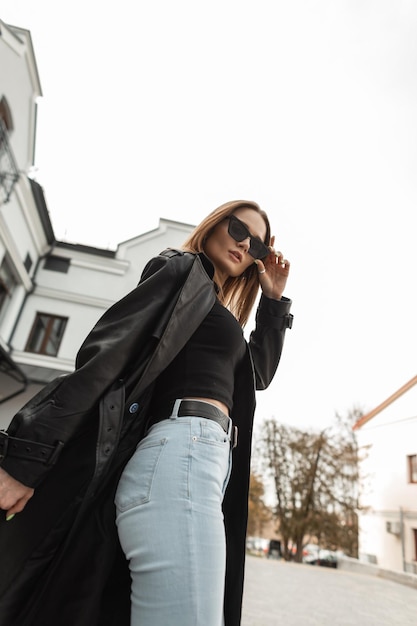 Stijlvolle mooie jonge vrouw in een modieuze zwarte leren lange jas met jeans met hoge taille en een tanktop repareert een coole zonnebril in de stad Prachtige vrouwelijke jurk in stedelijke stijl