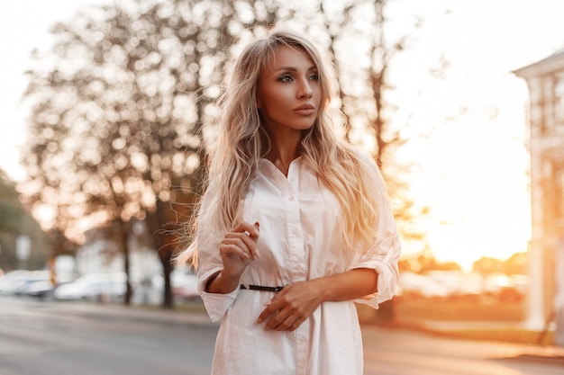 Stijlvolle mooie jonge vrouw in modieus wit overhemd loopt op straat bij zonsondergang