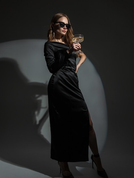 Stijlvolle mooie jonge vrouw met coole zonnebril in een mode zwarte elegante jurk met hakken houdt een glas champagne vast op een feestje Lady viert een evenement