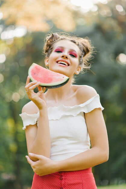 Stijlvolle mooie jonge vrouw met lichte, sappige make-up in de vorm van een watermeloen heeft een stuk watermeloen in haar handen