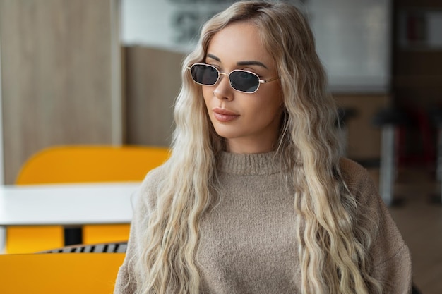 Stijlvolle mooie modieuze vrouw model met blond krullend kapsel met coole vintage zonnebril in een mode trendy beige trui zit in een café