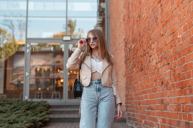 Stijlvolle mooie trendy vrouw in een modieuze leren jas met vintage hoge taille jeans en een tas draagt een zonnebril en loopt in de buurt van het winkelcentrum in de stad