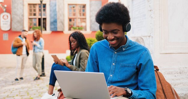 Stijlvolle multiraciale student die zijn laptopcomputer gebruikt, terwijl hij geniet op straat. portret van tiener afro-amerikaanse man buiten in de zomer. stedelijke achtergrond. jeugd concept