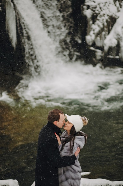 Foto stijlvolle paar verliefd zoenen in de winter besneeuwde bergen gelukkig romantische man en vrouw in luxe kleding zachtjes omhelzen bij waterval in de sneeuw vakantie uitje samen