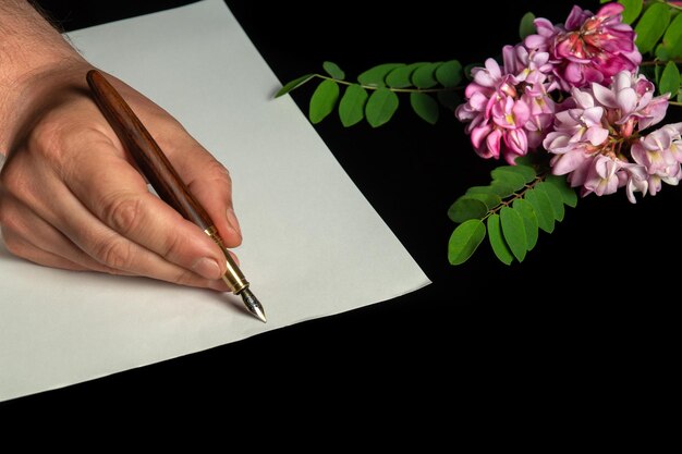 Foto stijlvolle pen in de hand van een man op een blanco vel papier en bloemen op de achtergrond. idee voor het schrijven van felicitaties voor een jubileum of feest