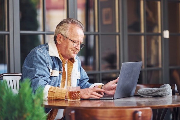 Stijlvolle senior in modieuze kleding en bril zit in het café en werkt met laptop.