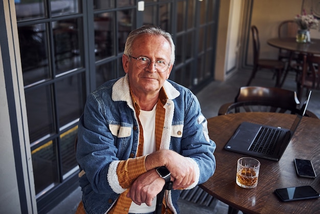 Stijlvolle senior in modieuze kleding en in glazen zit in het café met drankje en laptop op tafel.