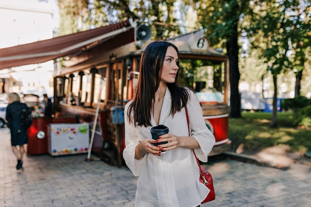 Stijlvolle spectaculaire dame met donker haar die een wit overhemd draagt, houdt een kopje koffie vast en kijkt opzij op de achtergrond van het stadspark in een goed humeur