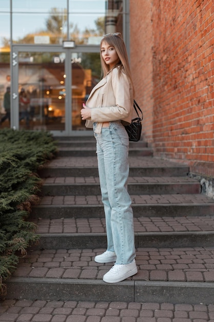 Stijlvolle straat mooie jonge vrouw in modieuze vrijetijdskleding met leren jas vintage highwaisted jeans witte schoenen met zwarte tas staat in de buurt van een rode bakstenen winkelcentrum