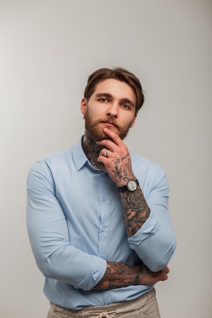 Stijlvolle succesvolle man hipster zakenman met baard en tatoeages in blauw fashion shirt en denken in studio op witte achtergrond