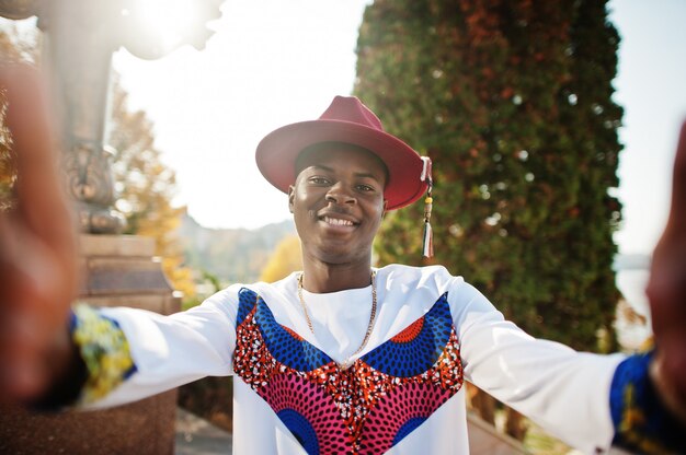 Stijlvolle trendy afro man in rode hoed en witte outfit gesteld op herfstdag.