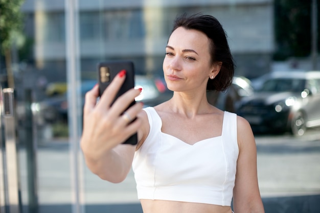 Stijlvolle volwassen vrouw in het wit neemt een selfie-portret aan de telefoon terwijl ze midden in een stedelijke straat staat tegen een achtergrond van glas