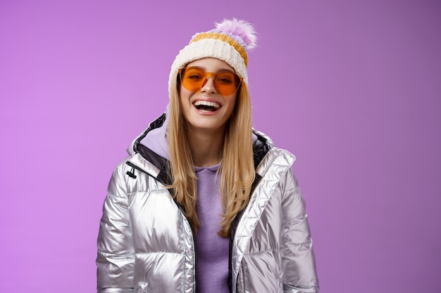 Stijlvolle vriendelijke charismatische blonde vrouw in zilveren glanzende jas hoed zonnebril klaar leren snowboarden glimlachen lachen gelukkig plezier besneeuwde recreatieoord, staande paarse achtergrond.