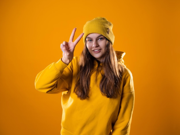 Stijlvolle vrolijke jonge brunette in een gele hoodie en hoeden toont een overwinningsgebaar met twee vingers...