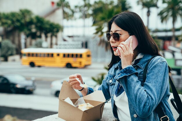 Stijlvolle vrouw die gebakken aardappel eet uit een afhaaldoos op de brug praat op mobiel. wazige achtergrond druk verkeer op straat, lokale dame in zonnebril met zakelijke telefoongesprekken tijdens de lunch.