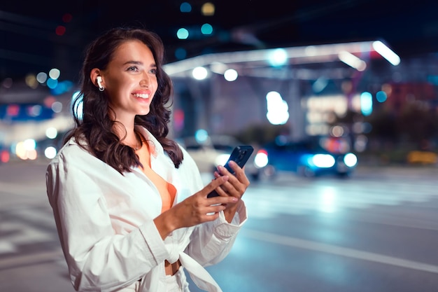 Stijlvolle vrouw die in de stad in de schemering op straat loopt met behulp van een smartphone en gratis muziek luistert