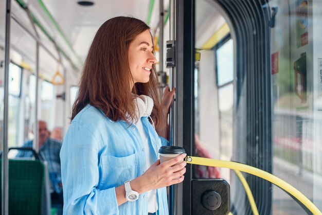 Stijlvolle vrouw in blauw hemd geniet van een reis in de moderne tram of bus staat met een kop koffie in de