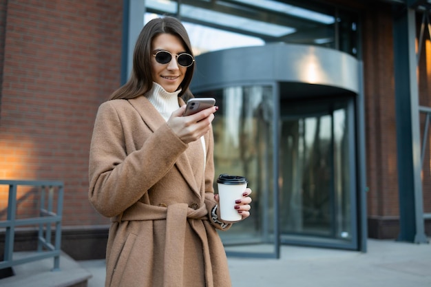 Stijlvolle vrouw in bruine jas en bril met koffiekopje met smartphone