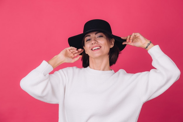 Stijlvolle vrouw in casual witte trui en hoed op roze rode muur