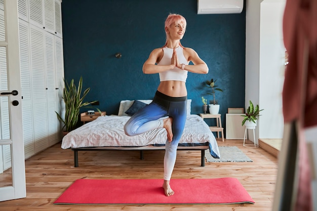 Stijlvolle vrouw in comfortabel trainingspak staat in vrksasana in de buurt van bed