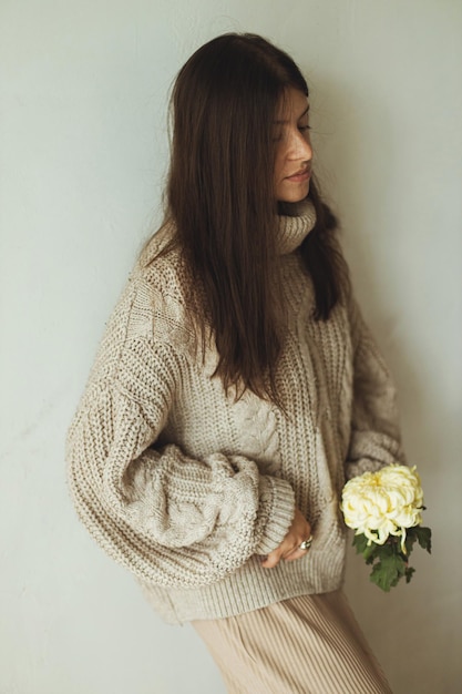 Stijlvolle vrouw in een gezellige gebreide trui die poseert met chrysanthemum op de achtergrond van een rustieke muur Gezellige herfstkleding Modieuze vrouw met bloemen humeurige herfst esthetiek