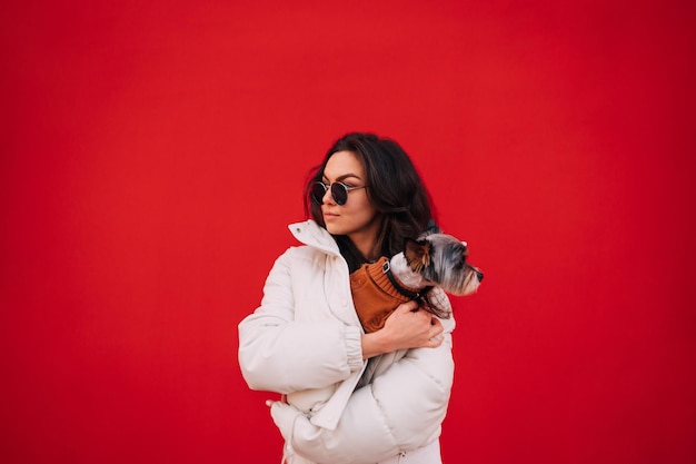 Stijlvolle vrouw in een jas staat op een achtergrond van een rode muur met een schattig hondje