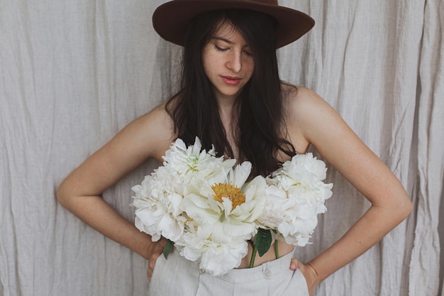 Stijlvolle vrouw in hoed poseren met witte pioenroos bloemen groeien uit broek Bloemige geur Zelfzorg