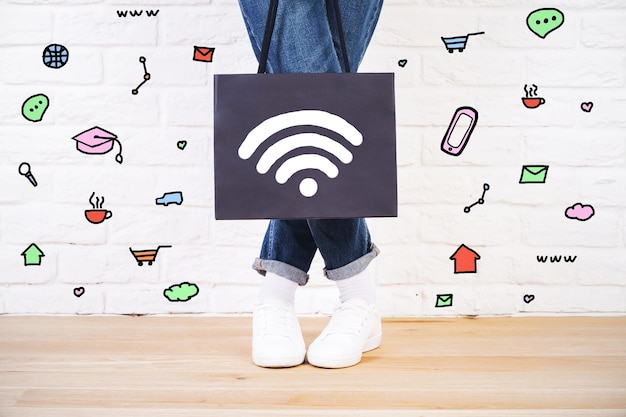 Foto stijlvolle vrouw in jeans en sneakers met boodschappentas met wifi-signaalpictogram. verschillende communicatiesymbolen rond op witte bakstenen muurachtergrond