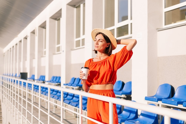 Stijlvolle vrouw in oranje kleding bij zonsondergang bij fietspad stadion poseren met kopje koffie