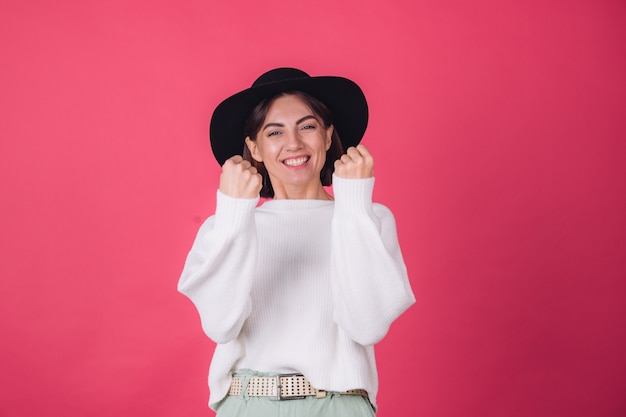 Stijlvolle vrouw in witte casual trui en hoed op rood roze muur