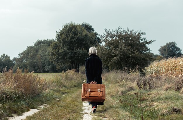 Stijlvolle vrouw in zwarte jas en koffer die op plattelandsweg loopt