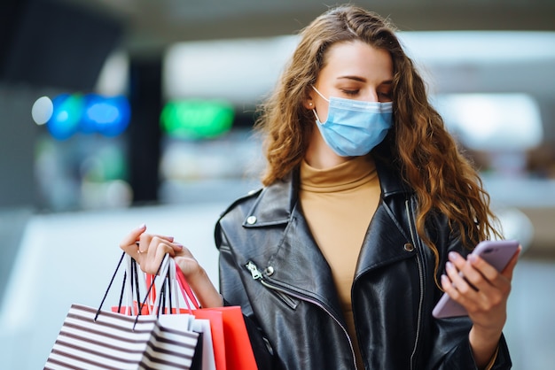 Stijlvolle vrouw met beschermend medisch masker met boodschappentassen die haar telefoon gebruikt. Mode, stijl, Black Friday,