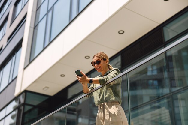 Stijlvolle vrouw ondernemer in zonnebril met behulp van telefoon terwijl staande op modern gebouw achtergrond