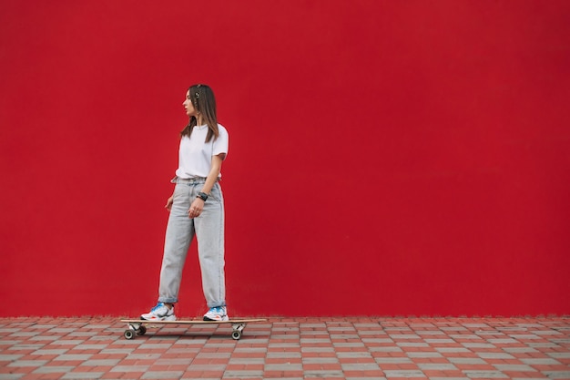 Stijlvolle vrouw rijdt op een longboard op straat op een achtergrond van een rode muur en kijkt naar de zijkant