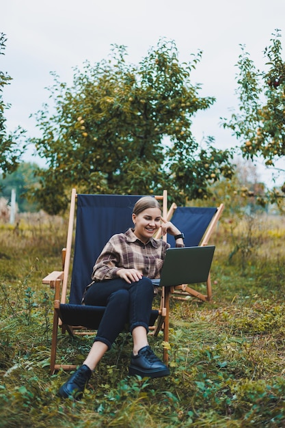 Stijlvolle vrouw zittend op een stoel in de buurt van groene planten in de tuin en op internet surfen op een laptop terwijl ze naar de zijkant kijkt Werk op afstand vrouwelijke freelancer